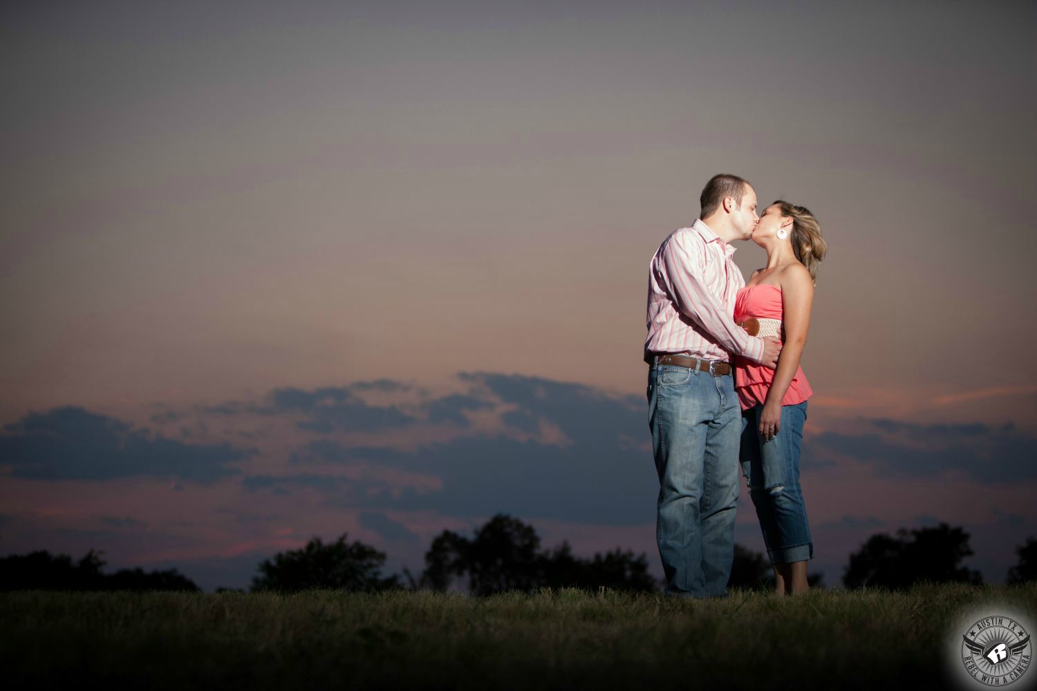 dirty blond girl with hoop earrings wearing a pink strapless top with a white belt, blue Capri jeans kissing a light brown haired guy in a white and pink striped dress shirt, brown belt and blue jeans standing on a slight rise in front of a grey and pink big Texas sky with a string of clouds in this tender engagement photo session in Round Rock, a suburb of Austin.   